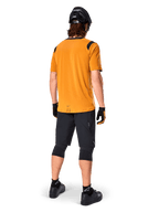 Alpinestars Maglia A-Dura Dri Wool a maniche corte, maglia da ciclismo, Dark Gold, vista posteriore di un mountain biker che indossa una maglia giallo senape con inserti in rete nera sulle spalle, abbinata a pantaloncini neri, ginocchiere, casco nero e guanti, abbigliamento tecnico da ciclismo fuoristrada