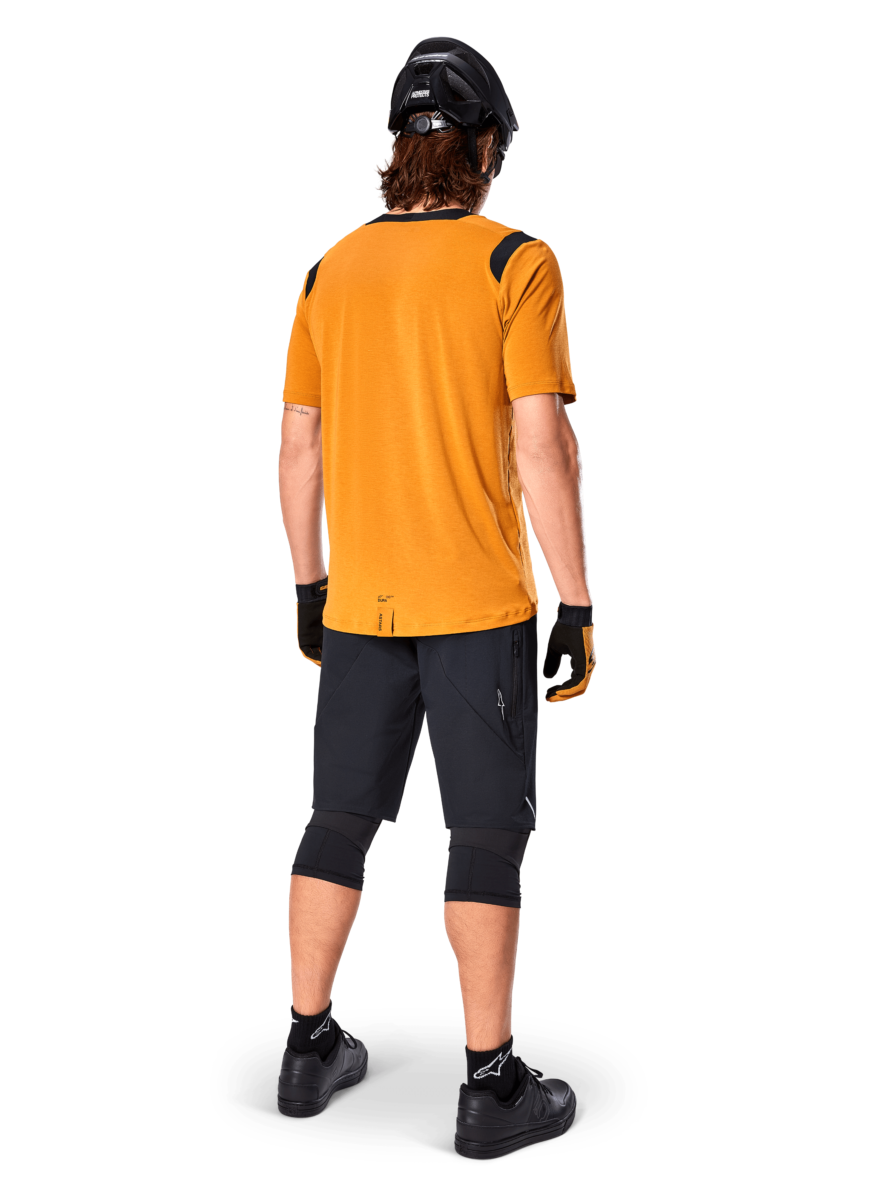 Alpinestars Maglia A-Dura Dri Wool a maniche corte, maglia da ciclismo, Dark Gold, vista posteriore di un mountain biker che indossa una maglia giallo senape con inserti in rete nera sulle spalle, abbinata a pantaloncini neri, ginocchiere, casco nero e guanti, abbigliamento tecnico da ciclismo fuoristrada