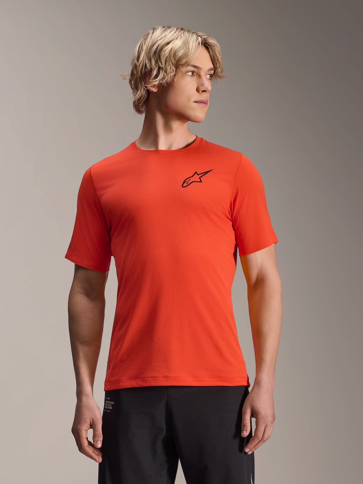 Alpinestars A-Aria Astar Short Sleeve Jersey, maglia da mountain bike MTB da uomo, arancione Spicy Orange con logo a stella nero, vestibilità performance, indossata da un modello biondo con pantaloni neri