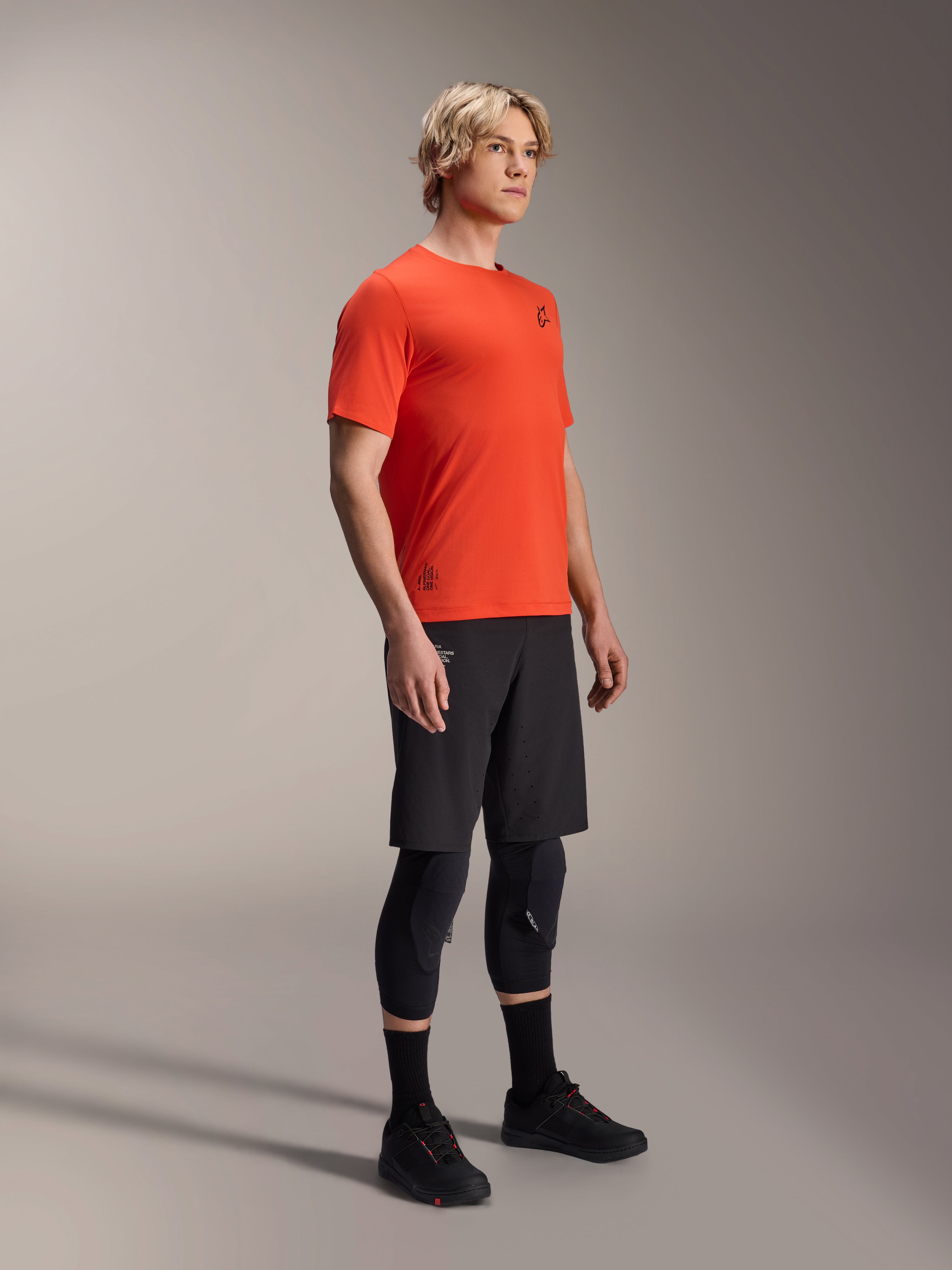 Maglia a maniche corte Alpinestars A-Aria Astar in Spicy Orange, indossata da un modello con pantaloncini MTB neri e ginocchiere, abbigliamento da mountain bike leggero e traspirante, arancione vivace con branding nero minimale