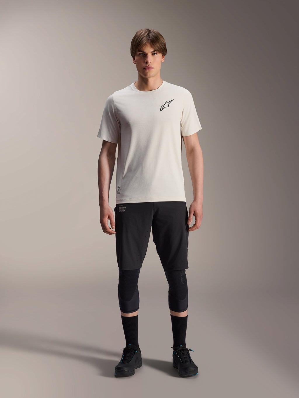 Modello che indossa la maglia a maniche corte Alpinestars A-Aria Astar in ecrù, abbinata a pantaloncini da MTB neri, ginocchiere e scarpe da ciclismo nere, caratterizzata da un logo a stella nero minimalista sul petto, progettata per il trail riding fuoristrada.