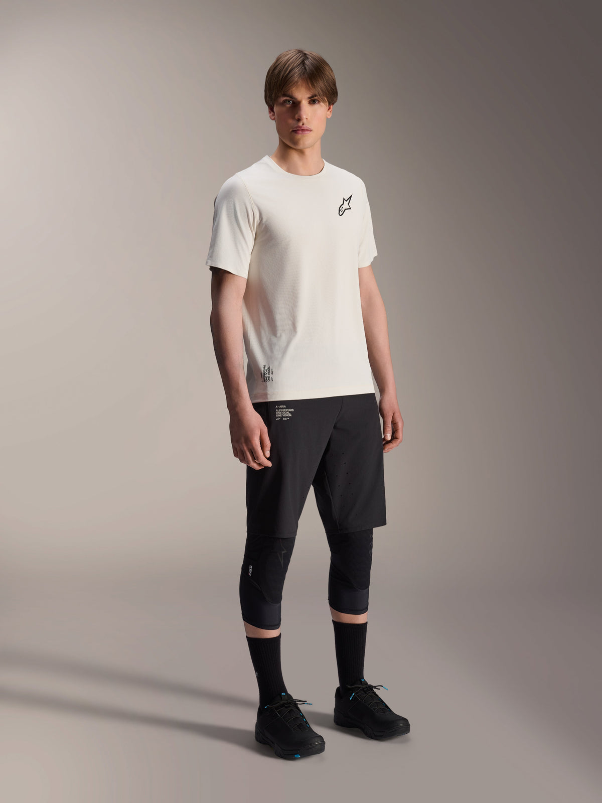 Alpinestars A-Aria Astar Jersey, a maniche corte, ecru, con logo nero sul petto, indossata con pantaloncini da ciclismo neri e ginocchiere, abbigliamento tecnico leggero per MTB