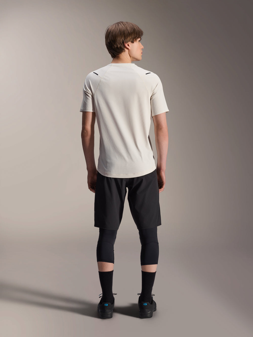 Modello maschile visto da dietro che indossa la maglia a maniche corte Alpinestars A-Aria Astar in colore Ecru, abbinata a pantaloncini da MTB neri e leggings protettivi, con coda più lunga e vestibilità performante per il trail riding fuoristrada