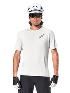 Ciclista che indossa la maglia a maniche corte Alpinestars A-Aria Astar, colore White Dove con piccolo logo Astar nero sul petto, casco da ciclismo bianco, occhiali da sole neri e guanti neri e bianchi, abbigliamento tecnico da MTB per fuoristrada