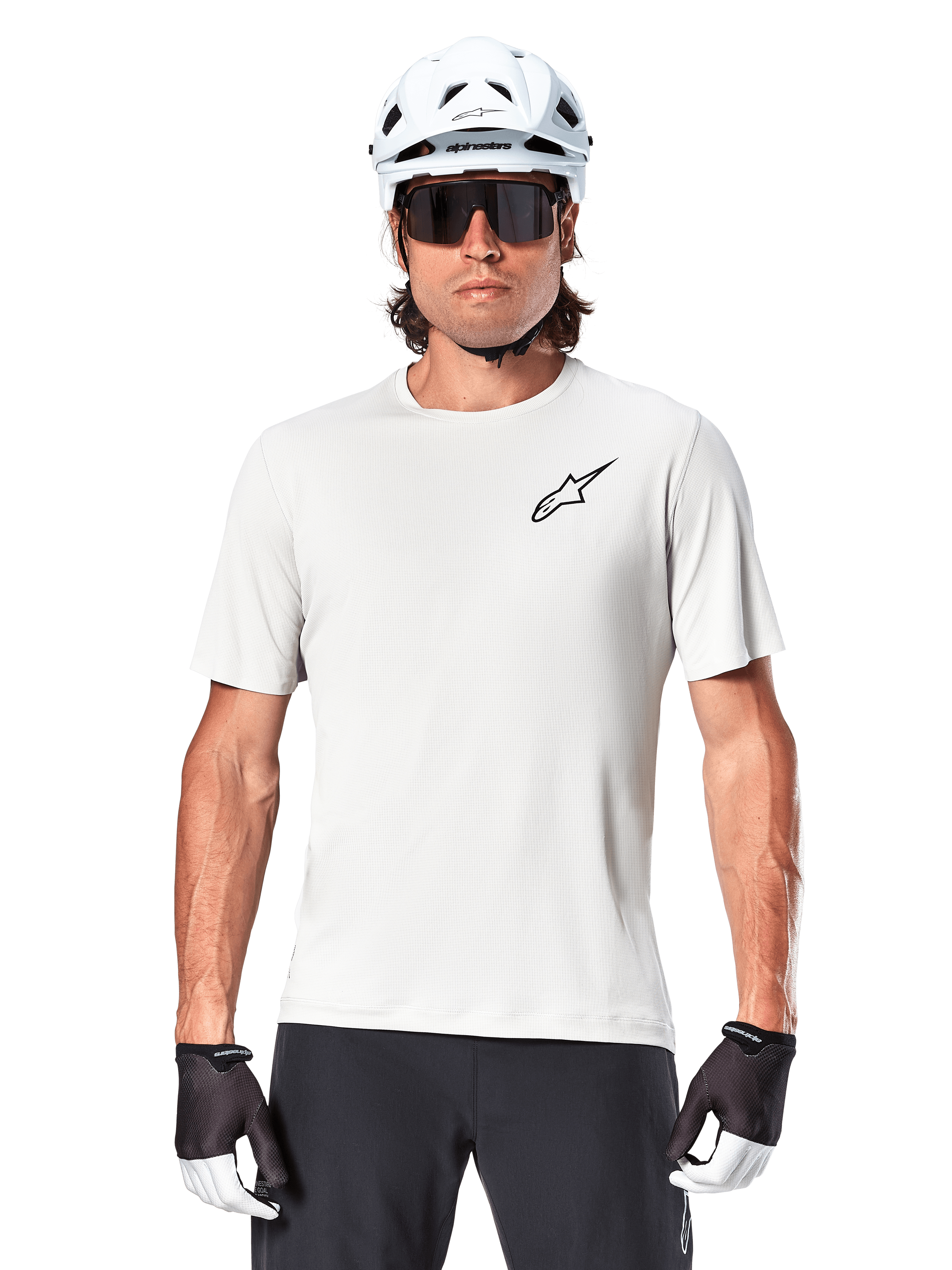 Ciclista che indossa la maglia a maniche corte Alpinestars A-Aria Astar, colore White Dove con piccolo logo Astar nero sul petto, casco da ciclismo bianco, occhiali da sole neri e guanti neri e bianchi, abbigliamento tecnico da MTB per fuoristrada