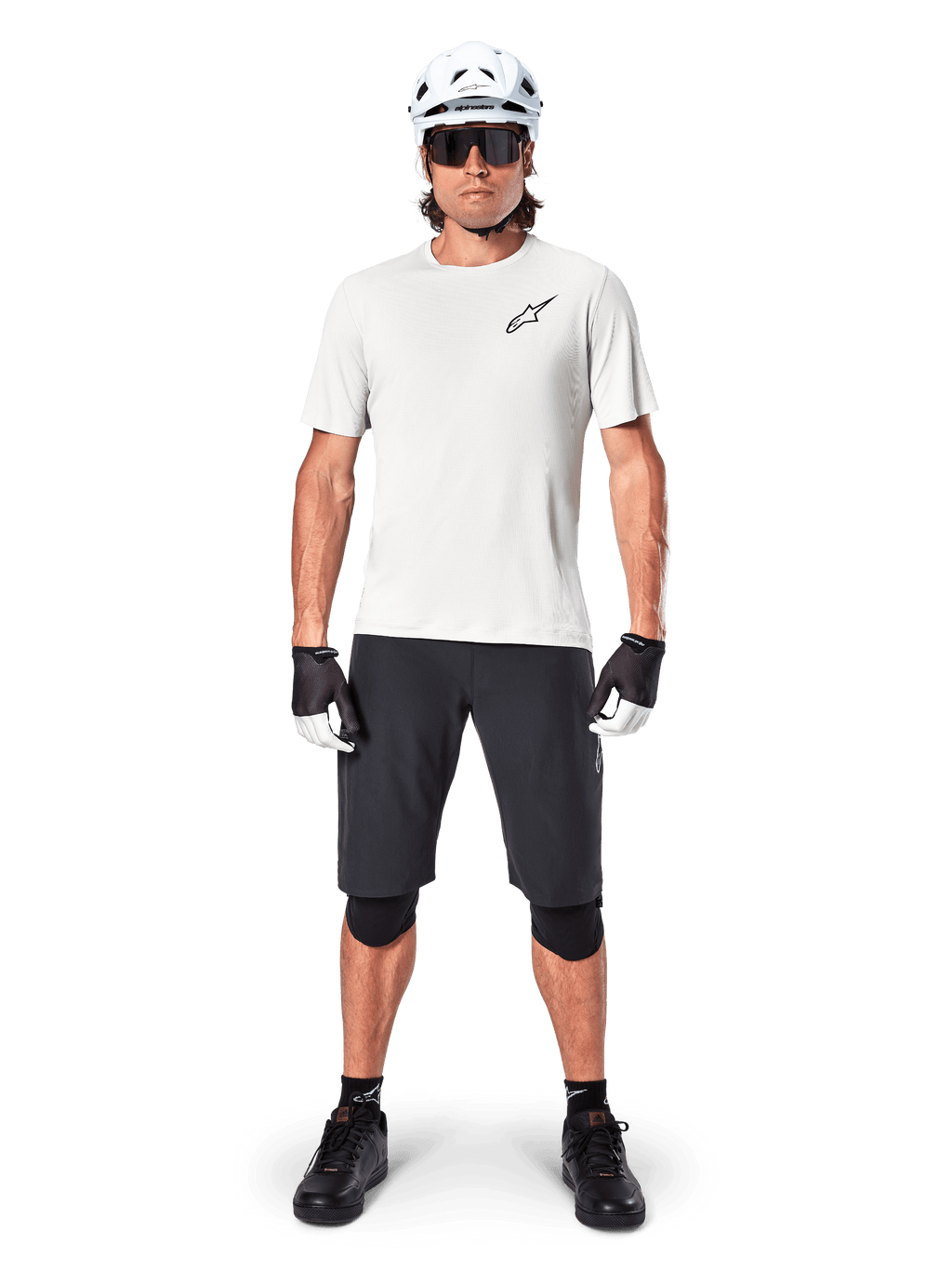 Ciclista uomo che indossa la maglia a maniche corte Alpinestars A-Aria Astar in White Dove, con logo Astars nero sul petto, abbinata a pantaloncini da mountain bike neri, casco bianco, occhiali da sole, ginocchiere nere e guanti, outfit completo da ciclismo fuoristrada.