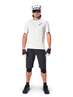 Ciclista uomo che indossa la maglia a maniche corte Alpinestars A-Aria Astar in White Dove, con logo Astars nero sul petto, abbinata a pantaloncini da mountain bike neri, casco bianco, occhiali da sole, ginocchiere nere e guanti, outfit completo da ciclismo fuoristrada.