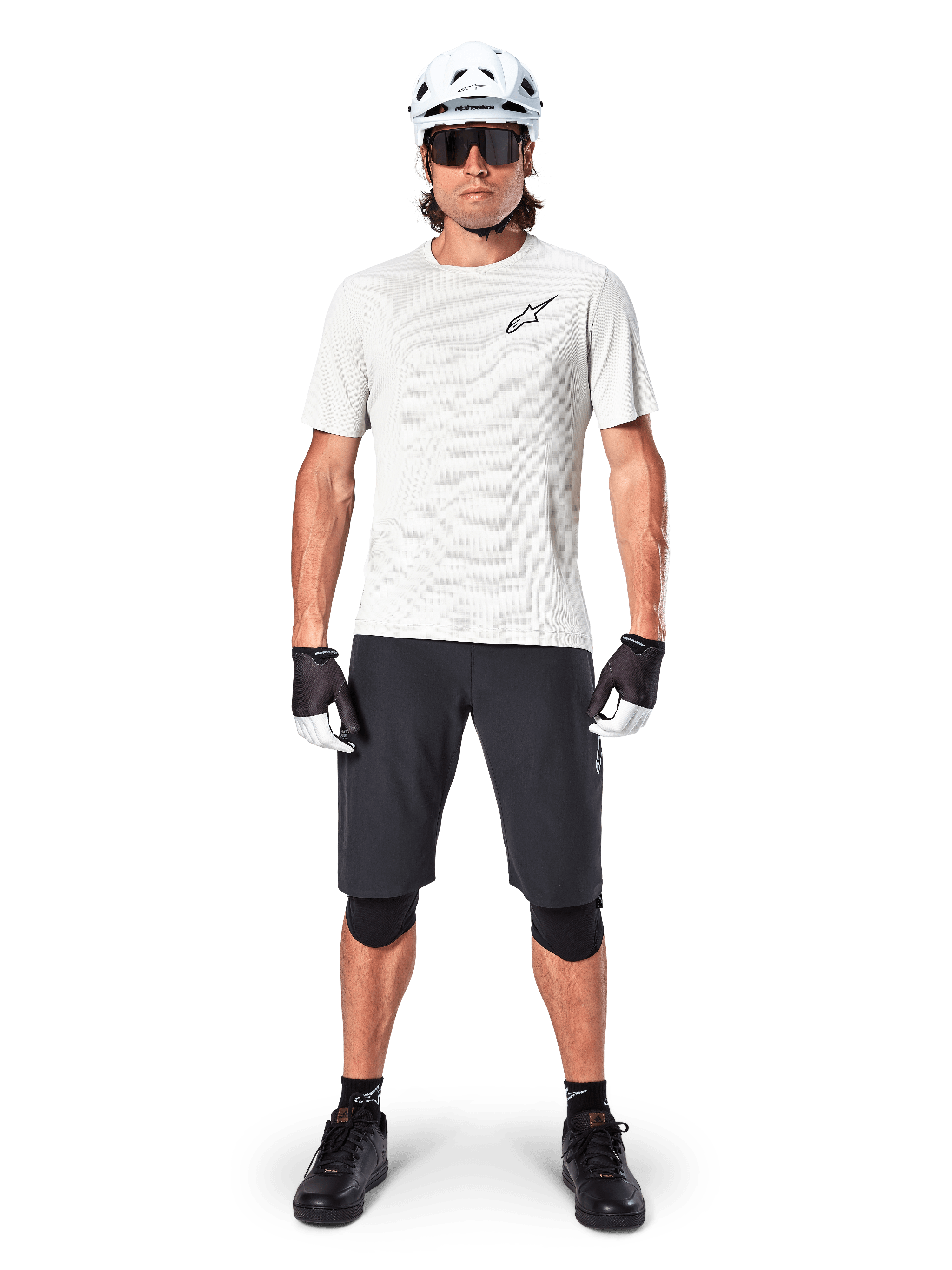 Ciclista uomo che indossa la maglia a maniche corte Alpinestars A-Aria Astar in White Dove, con logo Astars nero sul petto, abbinata a pantaloncini da mountain bike neri, casco bianco, occhiali da sole, ginocchiere nere e guanti, outfit completo da ciclismo fuoristrada.