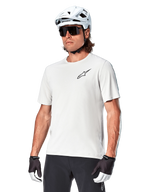 Alpinestars Maglia A-Aria Astar a maniche corte, White Dove, indossata da un ciclista con casco bianco e occhiali da sole scuri, con un piccolo logo a stella nero sul petto, abbigliamento performante traspirante per mountain bike