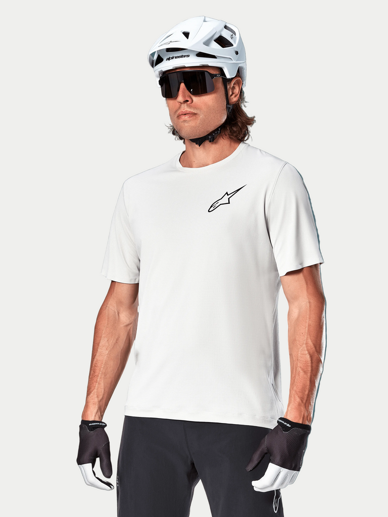 Alpinestars Maglia A-Aria Astar a maniche corte, White Dove, indossata da un ciclista con casco bianco e occhiali da sole scuri, con un piccolo logo a stella nero sul petto, abbigliamento performante traspirante per mountain bike