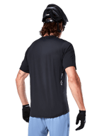 Alpinestars A-Dura Oscar Jersey, maglia da ciclismo a maniche corte, nero, vista posteriore su ciclista, abbinata a pantaloncini azzurri e casco MTB nero, piccolo logo bianco sul lato, abbigliamento tecnico per mountain bike