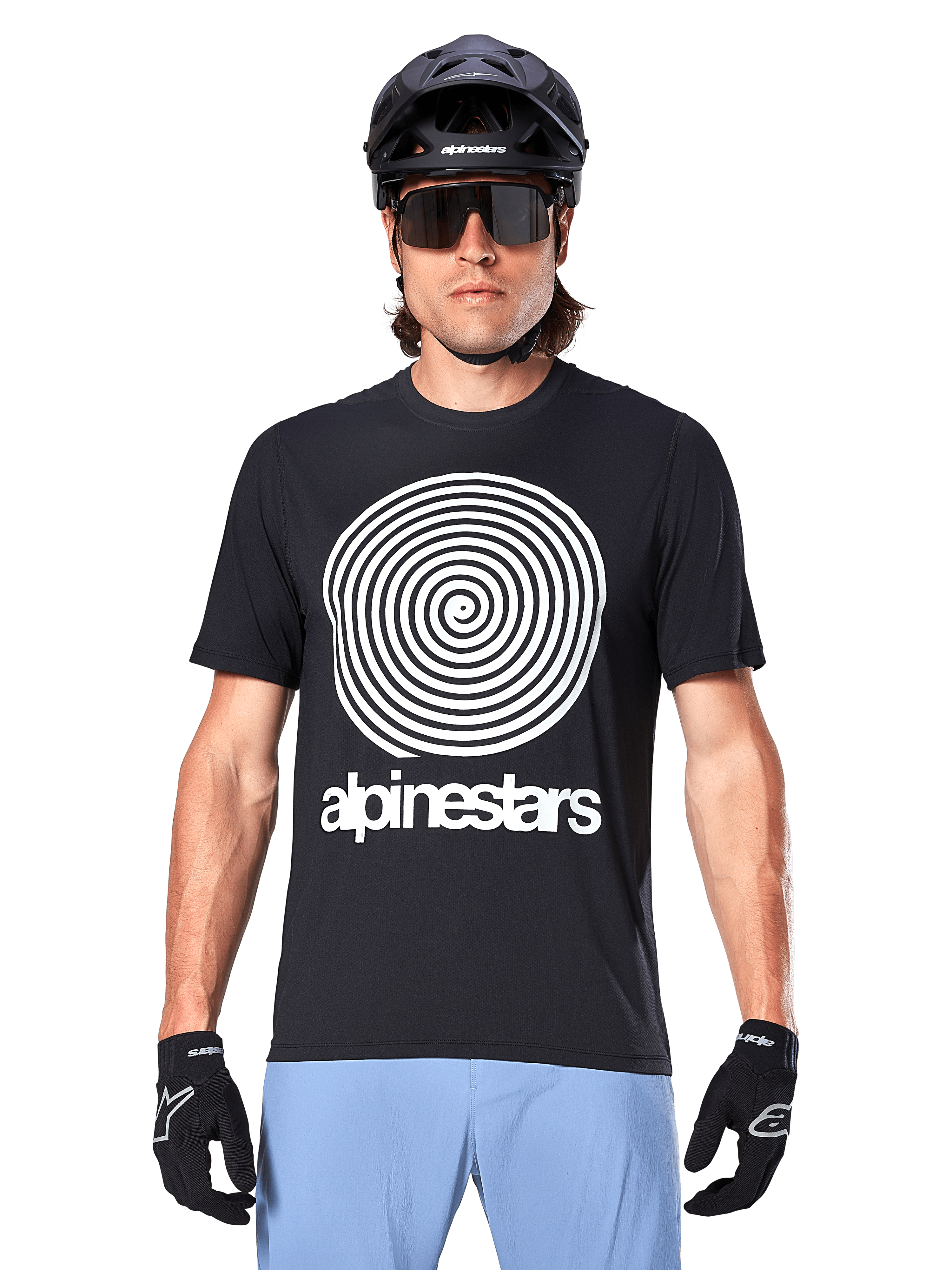 Alpinestars Maglia A-Dura Oscar, maglia MTB a maniche corte, nero, con una grande grafica a spirale vintage Oscar bianca sul petto, indossata con casco da ciclismo nero, occhiali da sole, guanti neri e pantaloncini azzurri, abbigliamento da ciclismo off-road d'ispirazione heritage