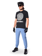Maglia a maniche corte Alpinestars A-Dura Oscar, maglia da MTB nera con grande grafica a spirale heritage bianca, indossata da un rider con casco nero, occhiali da sole, guanti neri e pantaloni azzurri, abbigliamento tecnico da mountain bike