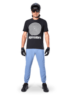 Alpinestars Maglia A-Dura Oscar a maniche corte, maglia da MTB nera con grande grafica a spirale Oscar vintage bianca e testo del brand, indossata con pantaloni azzurri, casco nero, occhiali da sole e guanti neri, abbigliamento tecnico da mountain bike
