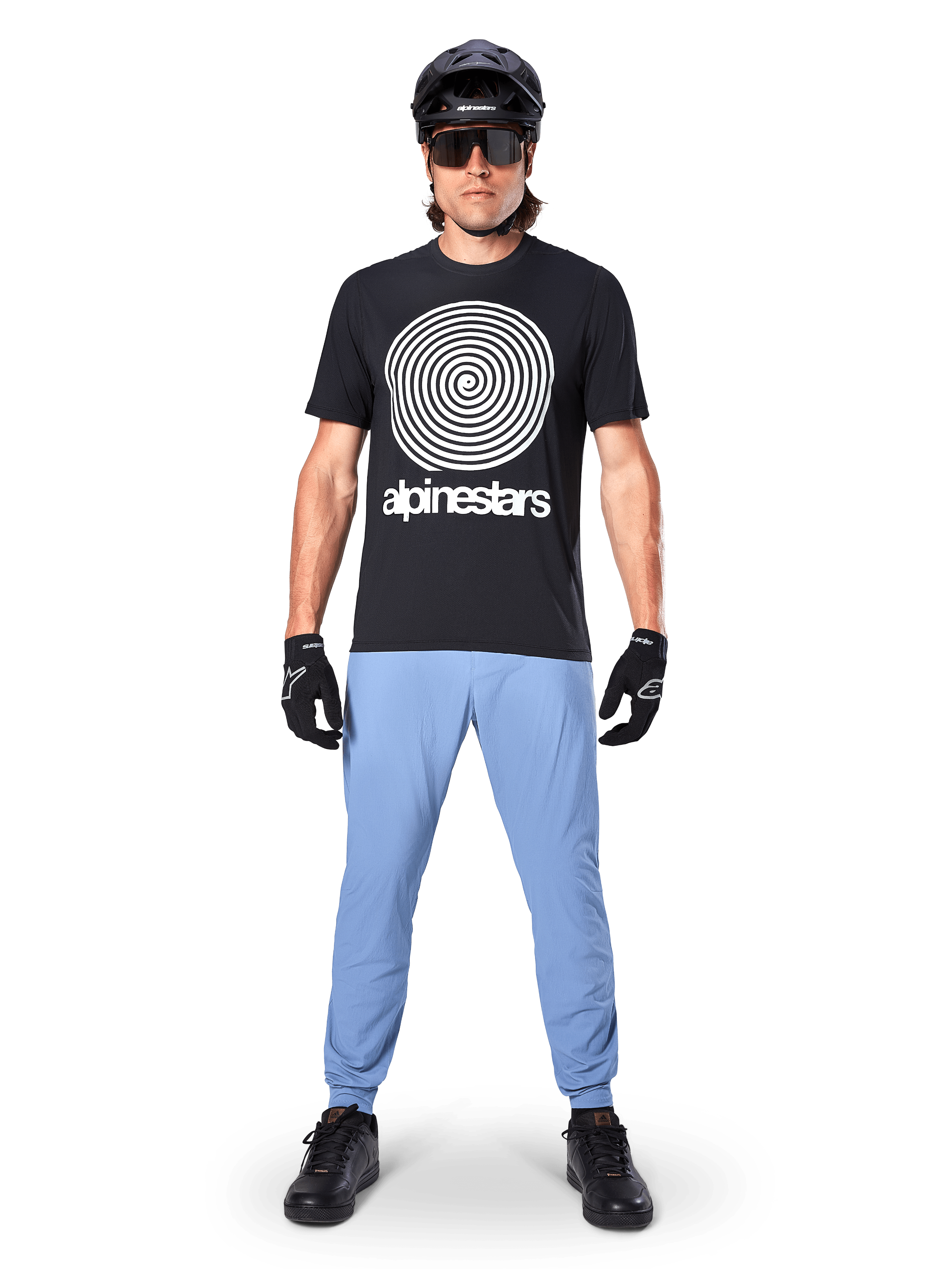 Alpinestars Maglia A-Dura Oscar a maniche corte, maglia da MTB nera con grande grafica a spirale Oscar vintage bianca e testo del brand, indossata con pantaloni azzurri, casco nero, occhiali da sole e guanti neri, abbigliamento tecnico da mountain bike