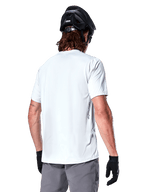 Alpinestars Maglia A-Dura Oscar, maglia MTB a maniche corte, White Dove, vista posteriore su un ciclista con casco da ciclismo nero, pantaloncini grigi e guanti neri, abbigliamento tecnico da ciclismo