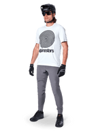 Alpinestars Maglia A-Dura Oscar a maniche corte, White Dove con grande grafica a spirale nera, indossata da un ciclista con pantaloni MTB grigi, casco nero e guanti, abbigliamento tecnico da ciclismo per guida off-road
