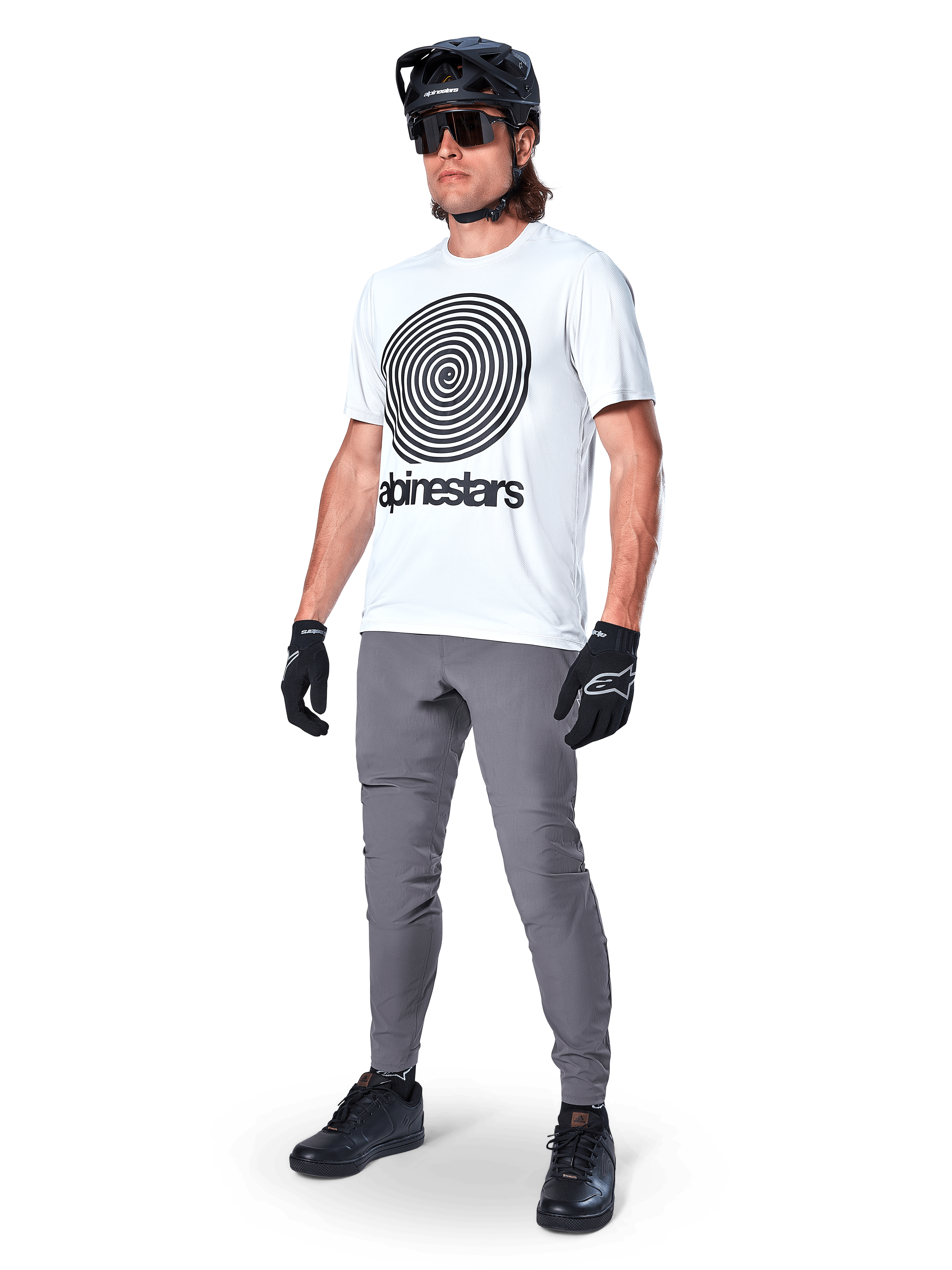 Alpinestars Maglia A-Dura Oscar a maniche corte, White Dove con grande grafica a spirale nera, indossata da un ciclista con pantaloni MTB grigi, casco nero e guanti, abbigliamento tecnico da ciclismo per guida off-road