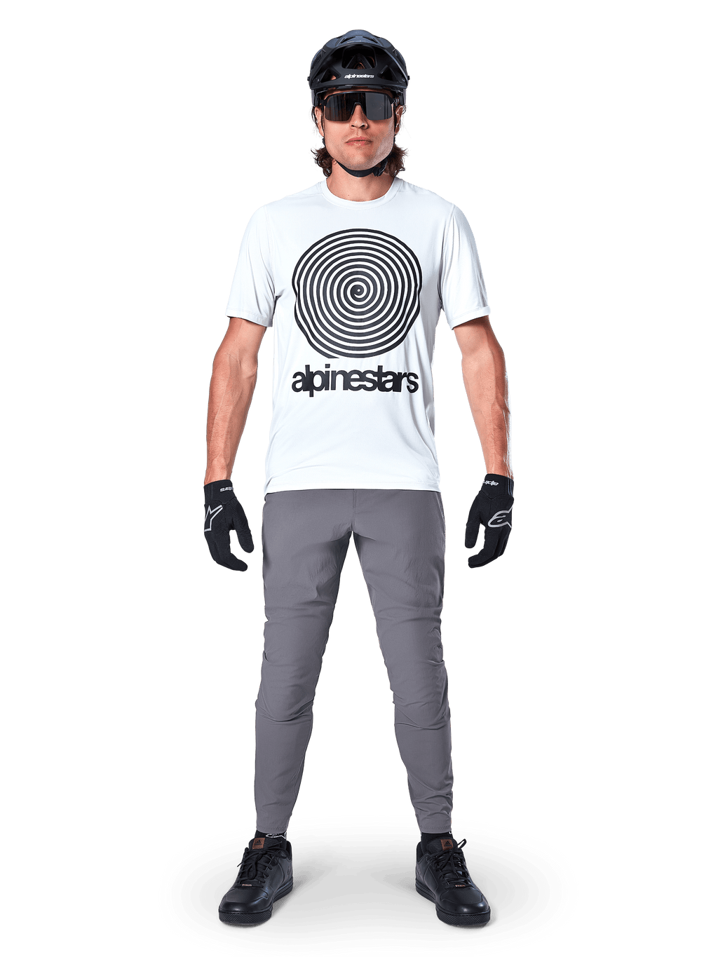 Mountain biker che indossa la maglia Alpinestars A-Dura Oscar a maniche corte, colore White Dove con grande logo a spirale nero, abbinata a pantaloni grigi, casco nero, occhiali da sole e guanti neri, abbigliamento tecnico per trail MTB