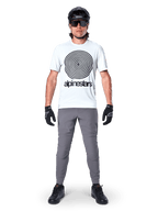 Mountain biker che indossa la maglia Alpinestars A-Dura Oscar a maniche corte, colore White Dove con grande logo a spirale nero, abbinata a pantaloni grigi, casco nero, occhiali da sole e guanti neri, abbigliamento tecnico per trail MTB
