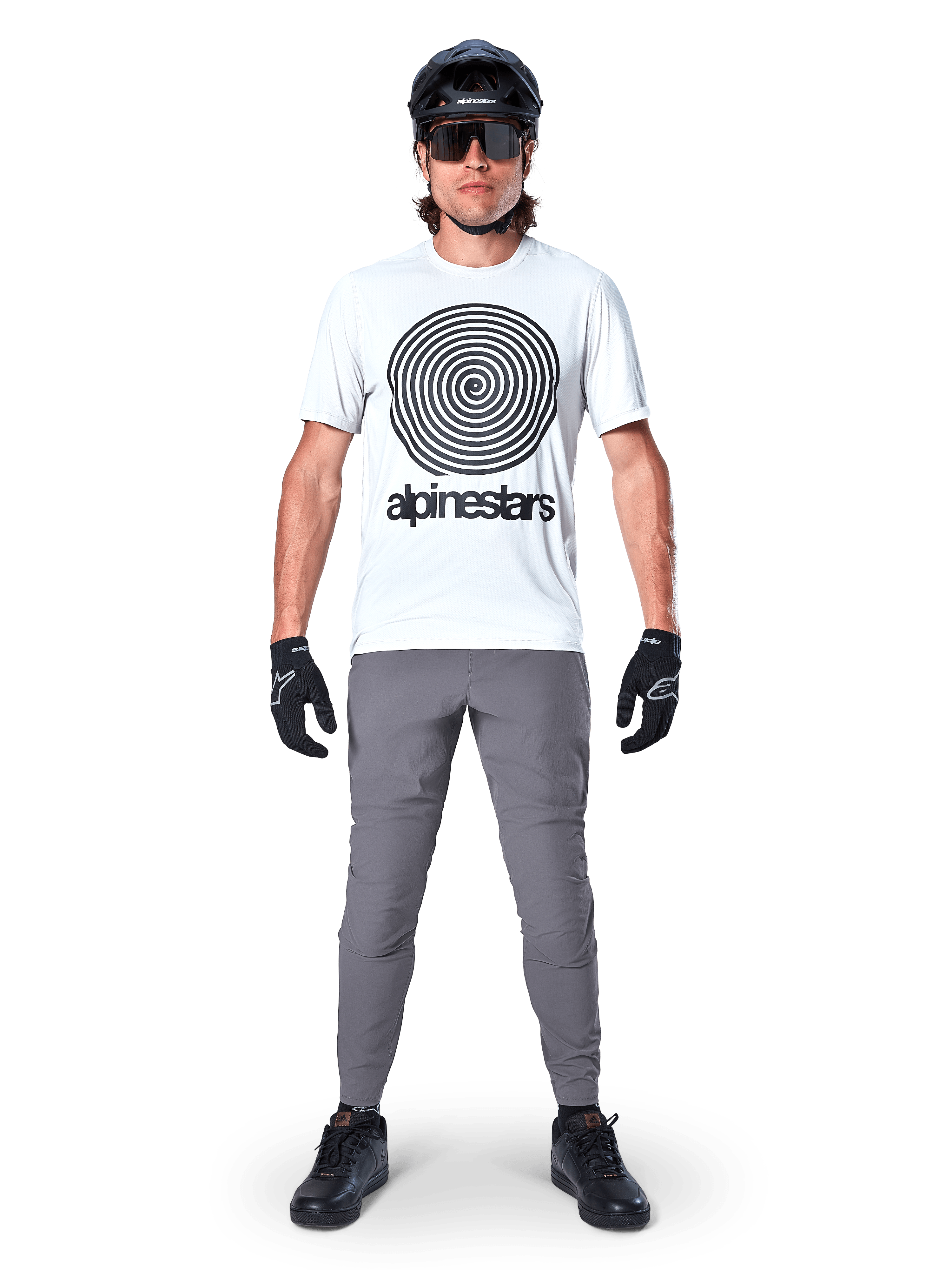 Mountain biker che indossa la maglia Alpinestars A-Dura Oscar a maniche corte, colore White Dove con grande logo a spirale nero, abbinata a pantaloni grigi, casco nero, occhiali da sole e guanti neri, abbigliamento tecnico per trail MTB