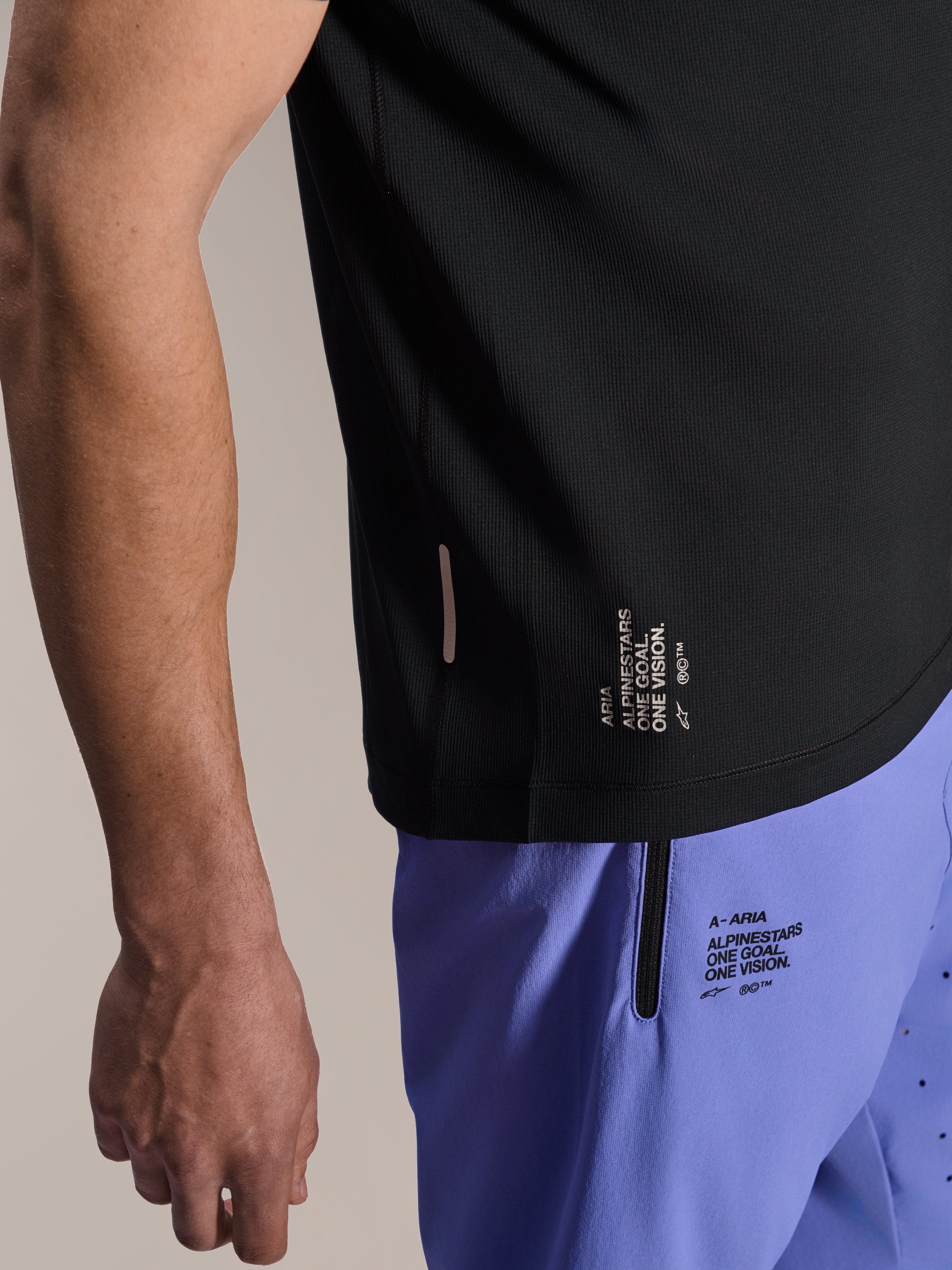 Alpinestars Maglia A-Aria Switch a maniche corte, maglia MTB nera, primo piano dell'orlo laterale con marchio bianco 'ONE GOAL. ONE VISION.', indossata con pantaloncini da ciclismo viola chiaro con loghi coordinati