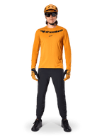 Alpinestars A-Dura Rocker Jersey - Long Sleeve in Dark Gold, con logo nero serigrafato sul petto, indossata con casco da MTB dorato coordinato, occhiali da sole e pantaloni neri, abbigliamento professionale da mountain bike