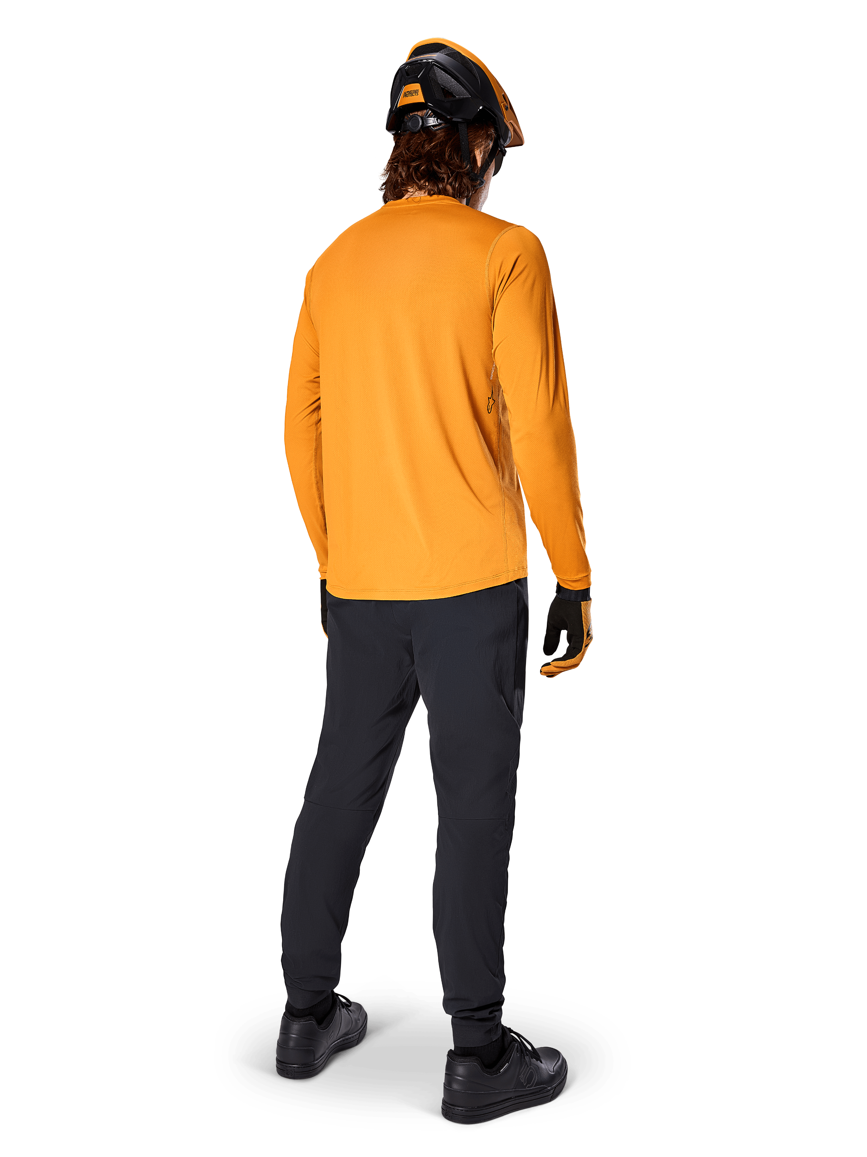 Ciclista di mountain bike visto da dietro che indossa la maglia a maniche lunghe Alpinestars A-Dura Rocker in Dark Gold, pantaloni neri, casco arancione e nero e scarpe nere, abbigliamento tecnico da MTB coordinato per trail riding