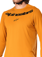 Alpinestars A-Dura Rocker Long Sleeve Jersey, oro scuro, vista ravvicinata del petto con scritta Alpinestars e logo a stella neri, tessuto jacquard testurizzato traspirante, abbigliamento tecnico da ciclismo MTB