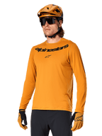 Alpinestars A-Dura Rocker Jersey a maniche lunghe, maglia da mountain bike MTB, Dark Gold, persona che indossa la maglia con casco e guanti coordinati, look sportivo coordinato con marchio Alpinestars sul petto