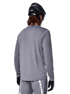 Maglia a maniche lunghe Alpinestars A-Dura Rocker in grigio scuro, vista posteriore di un ciclista di mountain bike, abbinata a pantaloncini grigi coordinati con branding bianco, casco da MTB nero e guanti neri, equipaggiamento professionale per ciclismo fuoristrada.