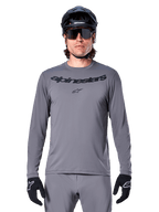Ciclista che indossa la maglia Alpinestars A-Dura Rocker a maniche lunghe, maglia da MTB grigio scuro con logo nero sul petto, abbinata a pantaloni grigi, casco nero e guanti, progettata per il ciclismo fuoristrada