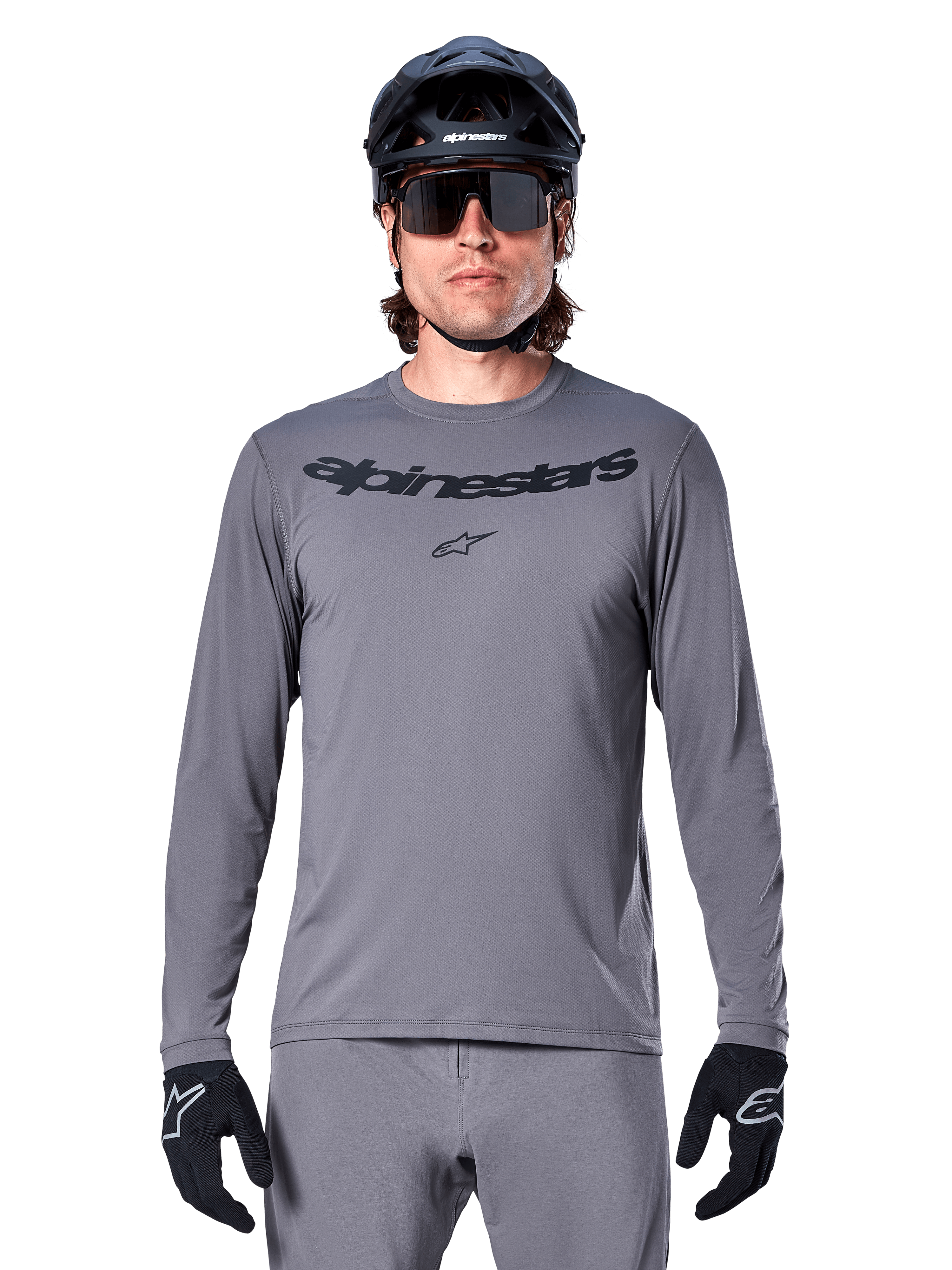 Ciclista che indossa la maglia Alpinestars A-Dura Rocker a maniche lunghe, maglia da MTB grigio scuro con logo nero sul petto, abbinata a pantaloni grigi, casco nero e guanti, progettata per il ciclismo fuoristrada