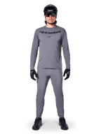 Ciclista che indossa la maglia a maniche lunghe Alpinestars A-Dura Rocker in grigio scuro, pantaloni grigi coordinati, casco da MTB nero, occhiali da sole e guanti, abbigliamento professionale da mountain bike per sentieri off-road