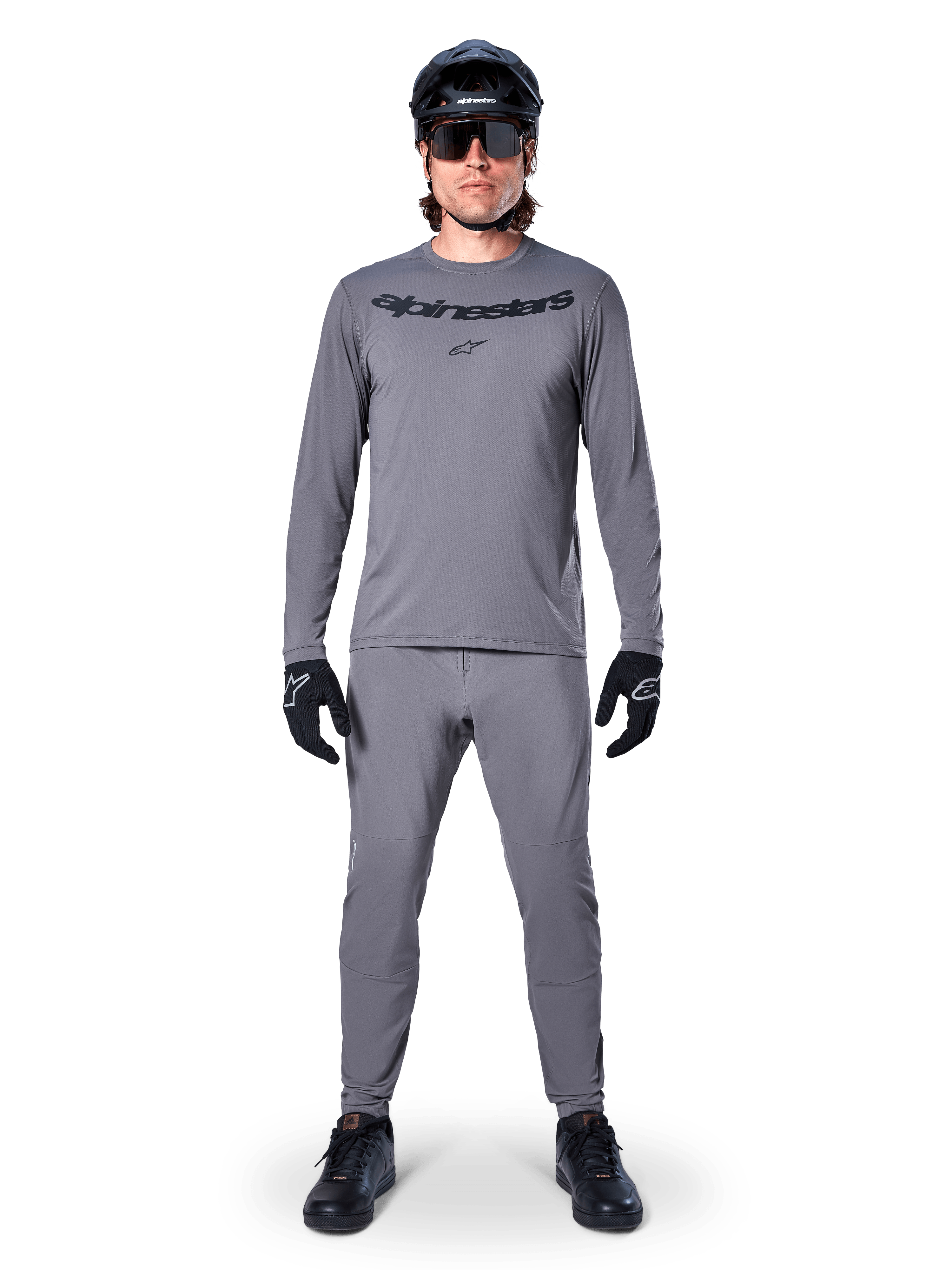 Ciclista che indossa la maglia a maniche lunghe Alpinestars A-Dura Rocker in grigio scuro, pantaloni grigi coordinati, casco da MTB nero, occhiali da sole e guanti, abbigliamento professionale da mountain bike per sentieri off-road