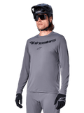Alpinestars A-Dura Rocker Jersey a maniche lunghe, maglia da mountain bike grigia, indossata con casco e guanti neri, con scritta nera sul petto, abbigliamento sportivo da ciclismo per il fuoristrada, sfondo neutro