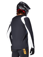 Ciclista MTB con maglia a maniche lunghe Alpinestars A-Supra Oscar, nero con pannelli bianchi sulle spalle e sulle maniche, vista posteriore, casco e guanti neri, pantaloni con accenti arancioni, abbigliamento da ciclismo fuoristrada resistente