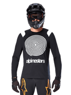 Maglia a maniche lunghe Alpinestars A-Supra Oscar, nera con grafica a spirale bianca e dettagli sulle spalle, indossata da un pilota con casco integrale nero e maschera, pantaloni neri e arancioni, abbigliamento tecnico da ciclismo per MTB gravity
