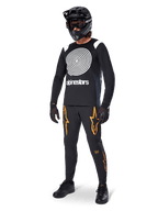 Pilota di MTB che indossa la maglia Alpinestars A-Supra Oscar a maniche lunghe nera, caratterizzata dalla grafica vintage a spirale Oscar bianca, pantaloni neri con loghi a stella arancioni, casco integrale nero con maschera e guanti neri, abbigliamento professionale da mountain bike