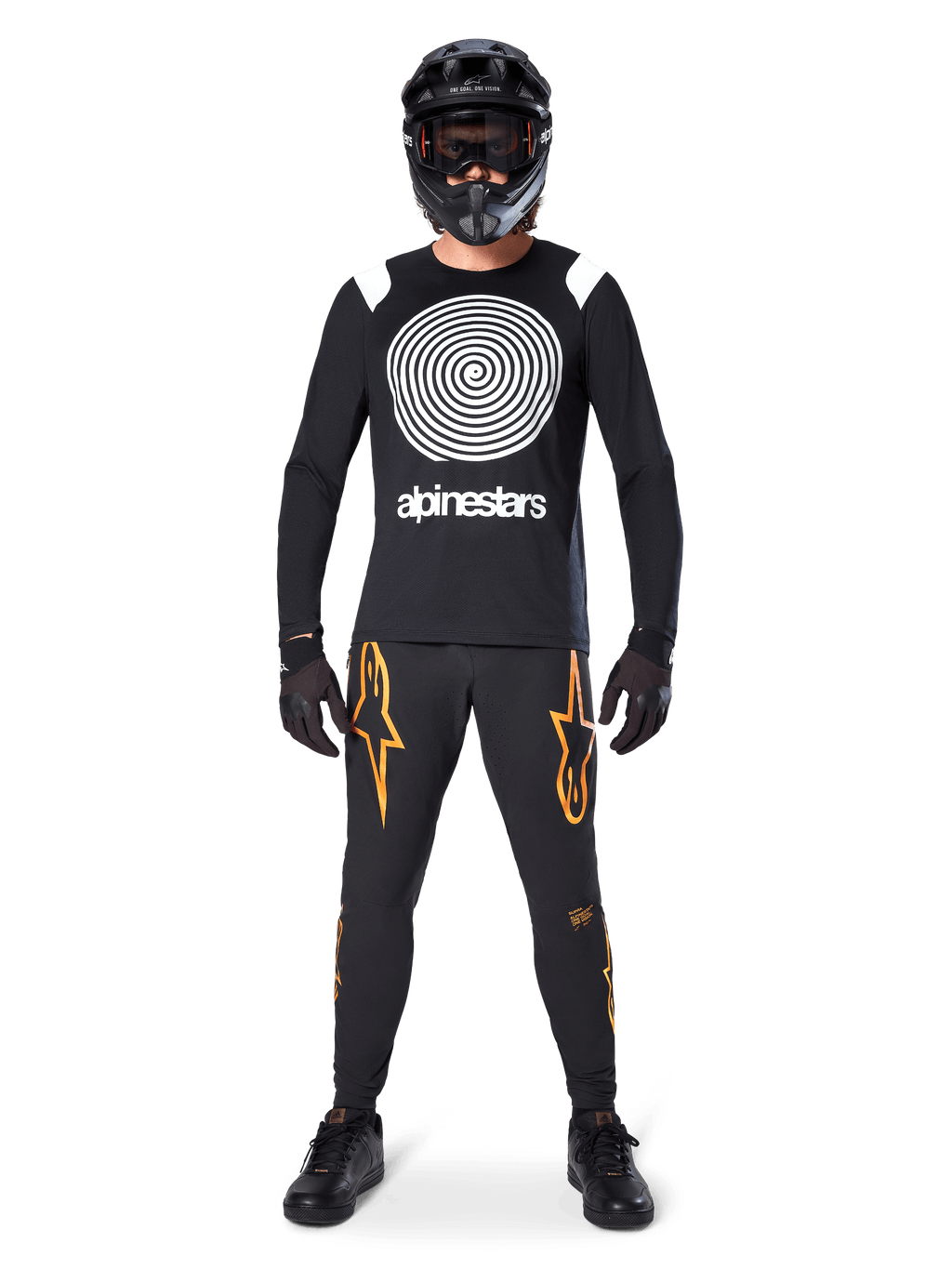 Modello con equipaggiamento completo da mountain bike, maglia a maniche lunghe Alpinestars A-Supra Oscar nera con grafica a spirale bianca sul petto, pantaloni neri con loghi a stella arancioni, casco integrale e maschera neri, guanti e scarpe neri, outfit coordinato per MTB gravity