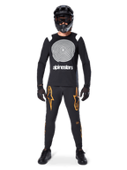 Modello con equipaggiamento completo da mountain bike, maglia a maniche lunghe Alpinestars A-Supra Oscar nera con grafica a spirale bianca sul petto, pantaloni neri con loghi a stella arancioni, casco integrale e maschera neri, guanti e scarpe neri, outfit coordinato per MTB gravity