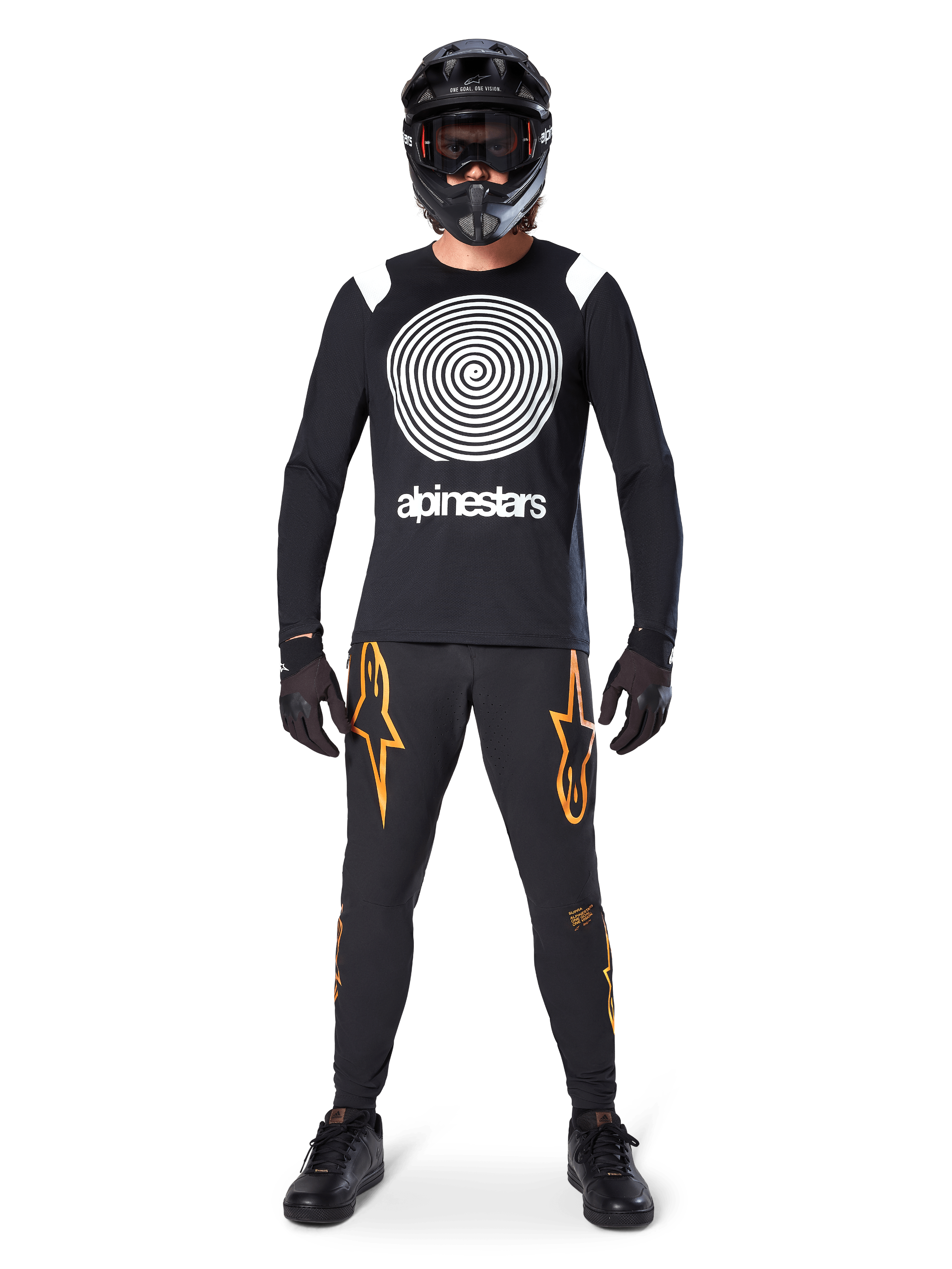 Modello con equipaggiamento completo da mountain bike, maglia a maniche lunghe Alpinestars A-Supra Oscar nera con grafica a spirale bianca sul petto, pantaloni neri con loghi a stella arancioni, casco integrale e maschera neri, guanti e scarpe neri, outfit coordinato per MTB gravity