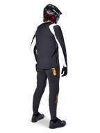 Maglia a maniche lunghe Alpinestars A-Supra Oscar, nera con pannelli bianchi sulle maniche, indossata da un rider MTB vista dal retro con casco integrale nero e pantaloni neri con loghi arancioni, abbigliamento da ciclismo per gravity off-road