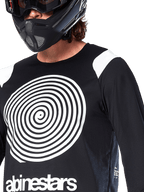 Alpinestars A-Supra Oscar Jersey, maglia a maniche lunghe, nero, con grande grafica a spirale Oscar bianca sul petto e logo del brand, indossata da un rider con casco integrale nero e maschera, abbigliamento tecnico da mountain bike per il gravity