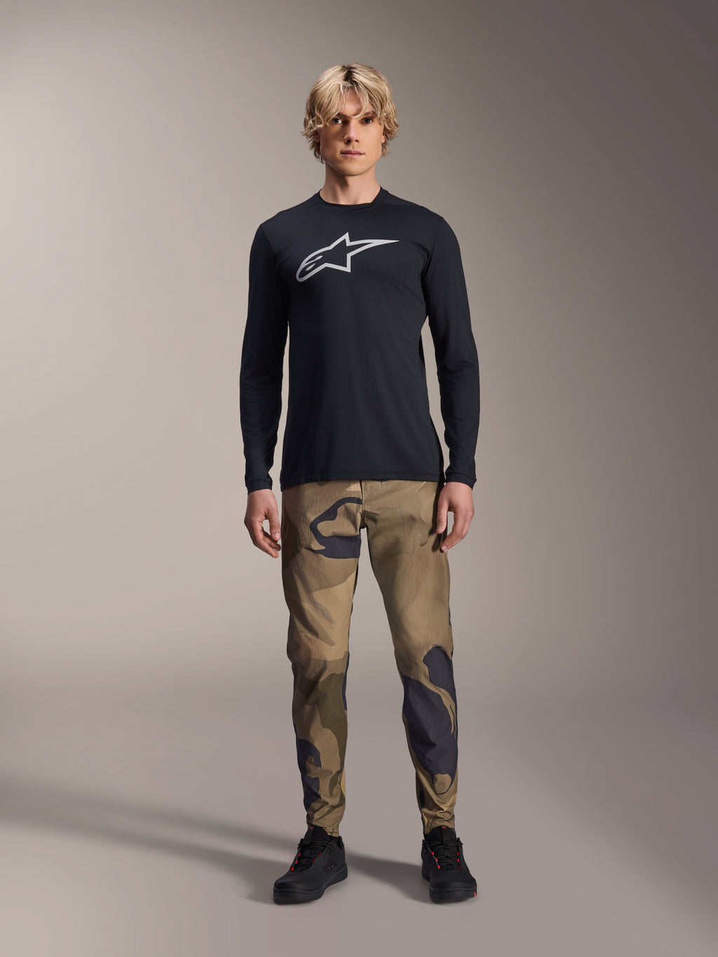 Maglia a maniche lunghe Alpinestars A-Dura Astar, nero, con un grande logo Astars bianco sul petto, indossata con pantaloni mimetici e scarpe nere, abbigliamento casual performante per mountain bike