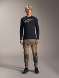 Alpinestars A-Dura Camo Pant, pantaloni da ciclismo MTB, camouflage marrone e verde, indossati con maglia nera a maniche lunghe e scarpe nere, stile sportivo da mountain bike con tessuto elasticizzato resistente