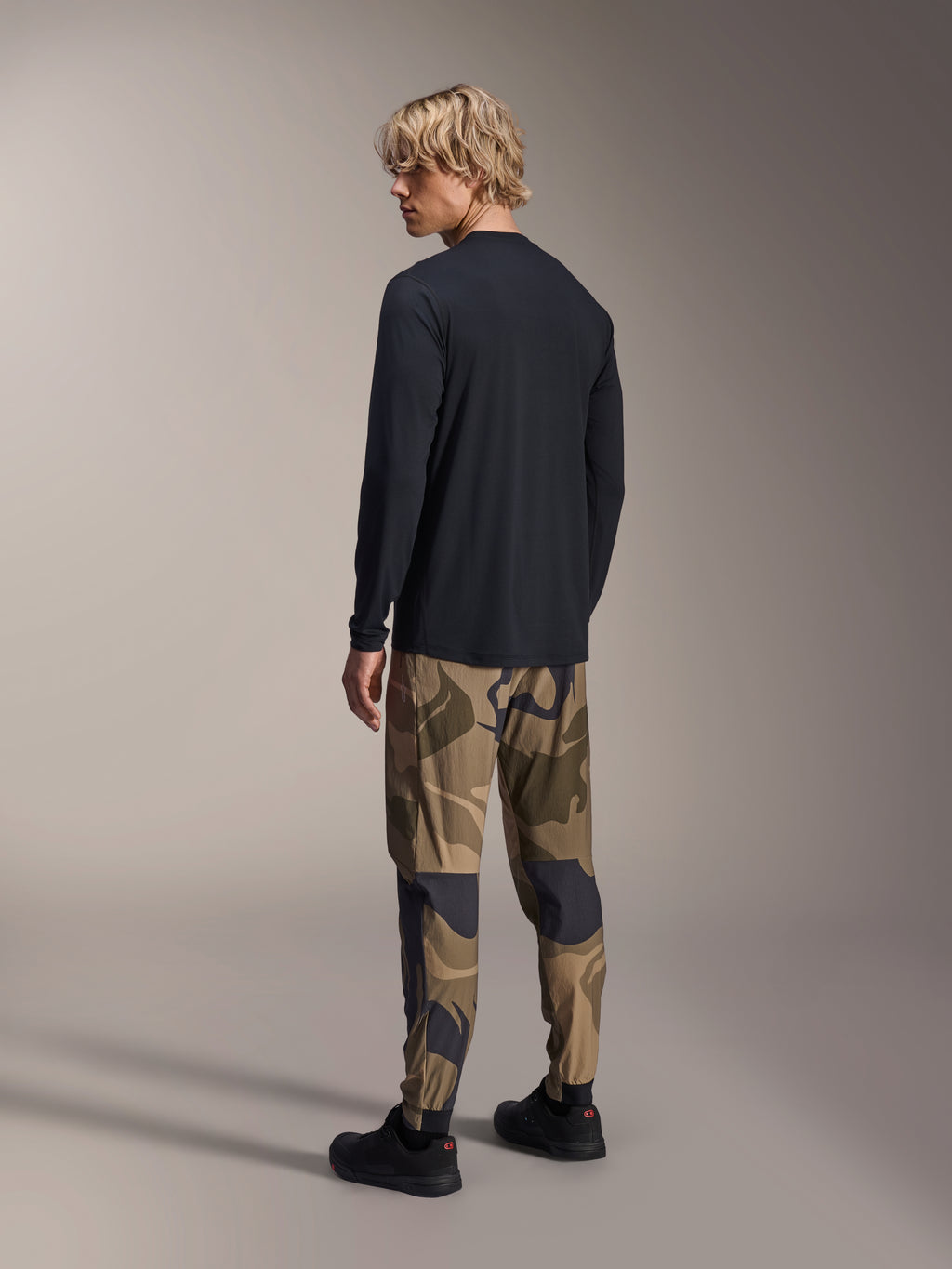 Alpinestars A-Dura Astar Long Sleeve Jersey, maglia MTB a maniche lunghe, nero, vista posteriore su modello, abbinata a pantaloni mimetici, abbigliamento tecnico casual con orlo posteriore allungato e vestibilità regolare