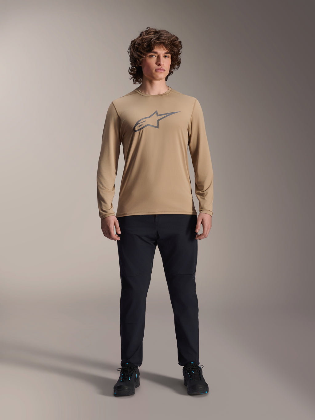 Alpinestars A-Dura Astar Long Sleeve Jersey, maglia a maniche lunghe marrone chiaro con logo Astars grigio sul petto, indossata da un modello con pantaloni tecnici e scarpe neri, abbigliamento tecnico casual per mountain bike