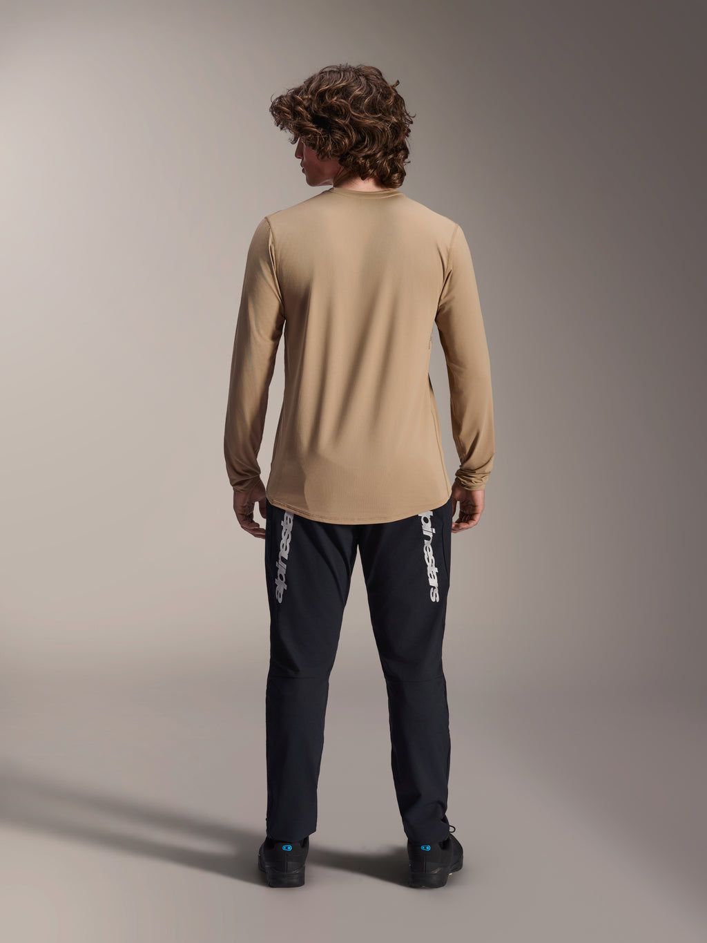 Maglia a maniche lunghe Alpinestars A-Dura Astar, marrone chiaro, vista posteriore che mostra la coda più lunga, indossata con pantaloni neri con marchio Alpinestars, abbigliamento tecnico da mountain bike