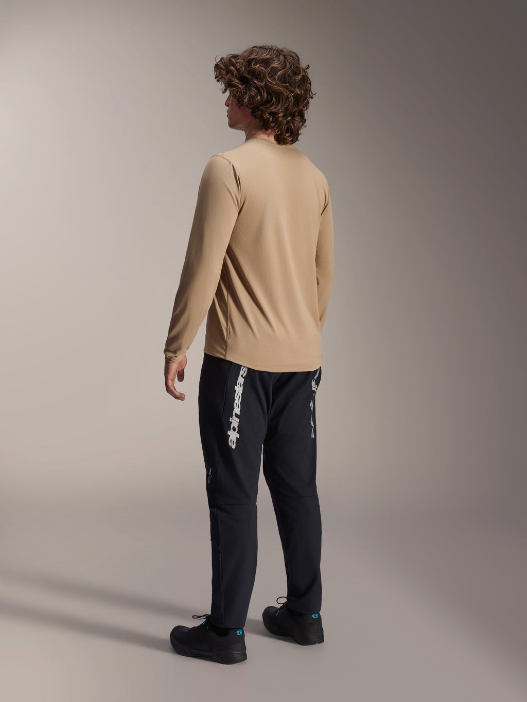 Maglia a maniche lunghe Alpinestars A-Dura Astar in marrone chiaro, vista posteriore di un modello maschile che mostra il design a coda allungata, abbinata a pantaloni e scarpe neri, abbigliamento tecnico casual per mountain bike.